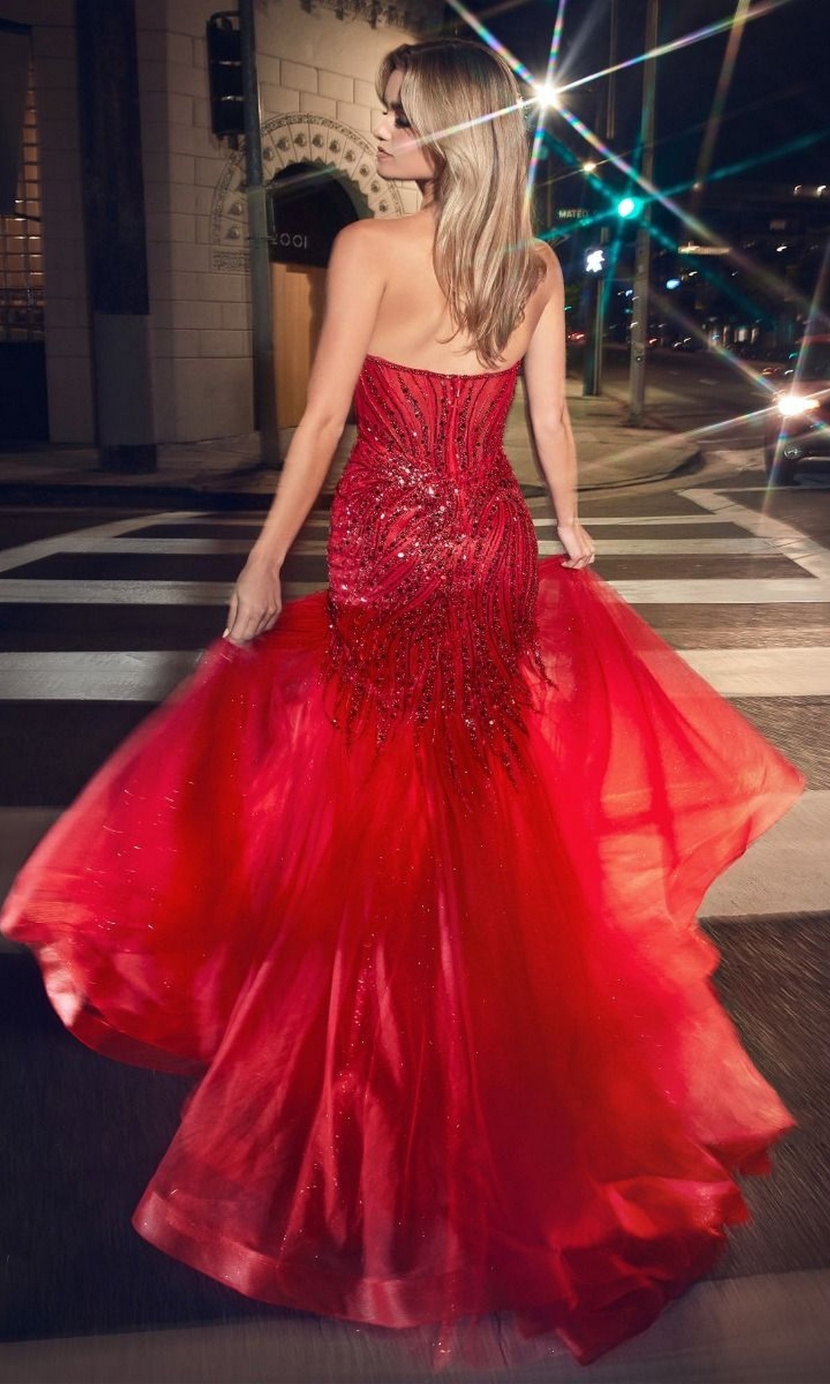 A woman wearing the Strapless Beaded Long Mermaid Prom Dress CD0214 stands on a city street at night, holding the flowing skirt of her gown and looking over her shoulder.